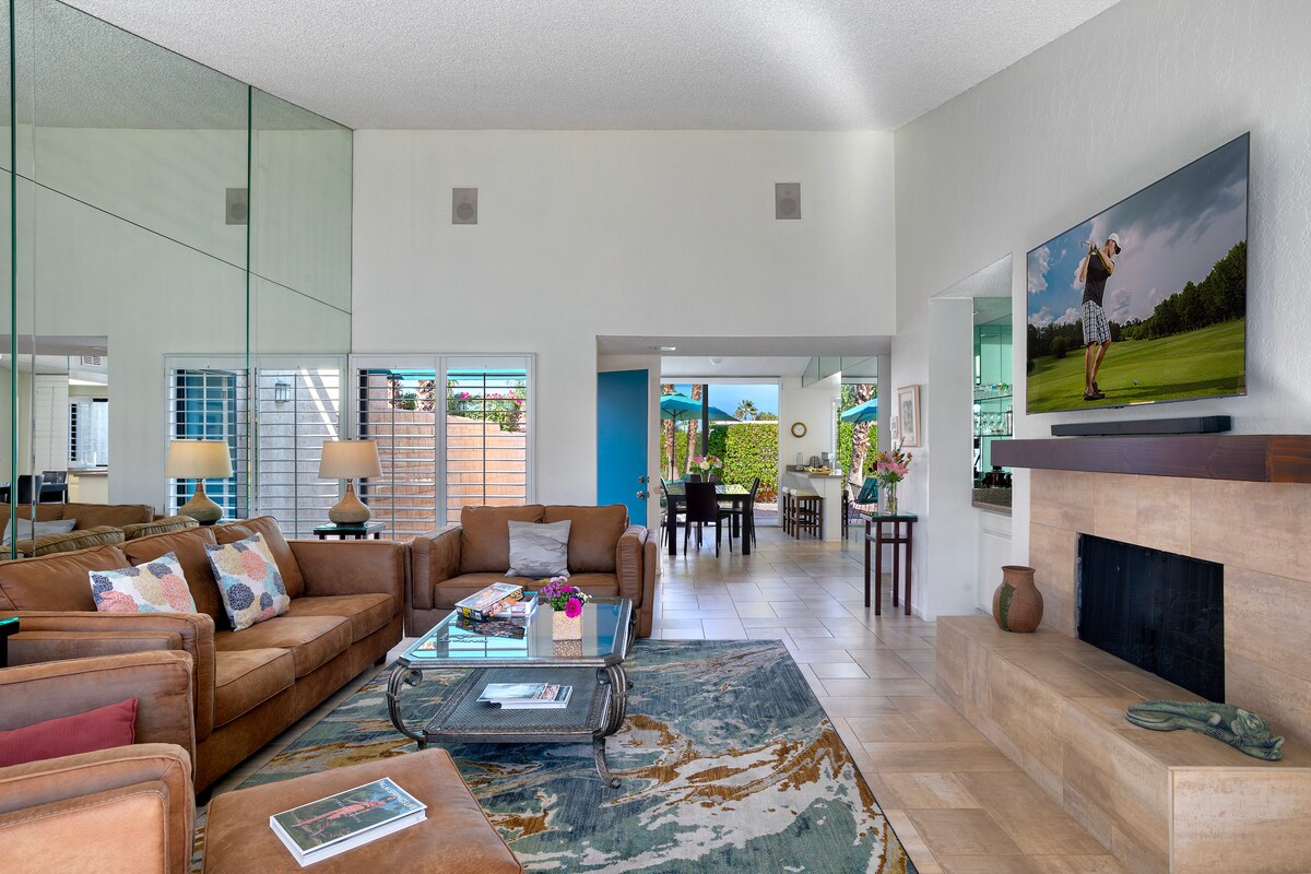 A spacious living area features a comfortable leather sofa, accent chairs, and a coffee table with decorative items. A large flat-screen TV is mounted on the wall beside a gas fireplace. Light enters through large windows, complementing the neutral color palette and open layout towards the dining area.