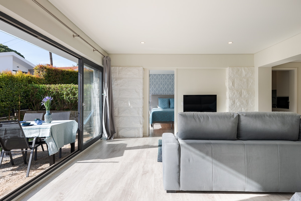 A spacious living area is showcased with large glass doors that open to an outdoor space. A comfortable gray sofa faces a wall-mounted television, while a dining table is visible through the glass doors, set for a meal with nature in the background.