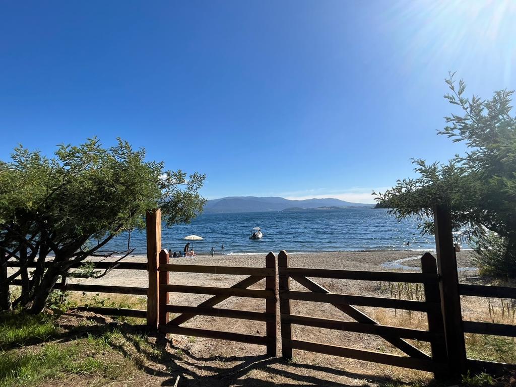 A scenic view of the beach is framed by wooden gates and lush greenery. The sandy shore is visible in the foreground, with gentle waves lapping at the edge. In the distance, a boat floats on the water, while a clear blue sky stretches overhead.