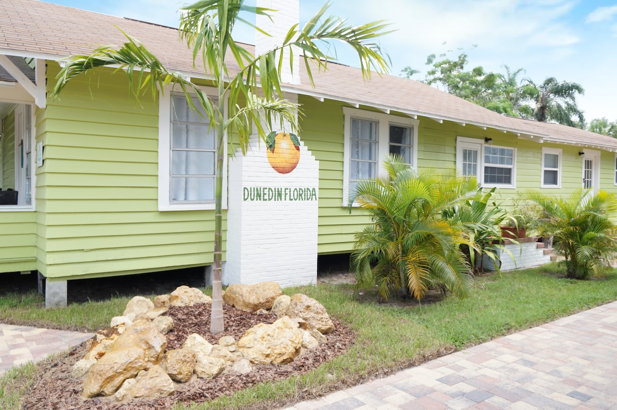 The exterior of a cozy duplex cottage is displayed, featuring a light green facade and a welcoming sign that reads 'Dunedin Florida.' Tropical plants and a decorative rock arrangement enhance the front yard, while a paved walkway leads to the entrance.