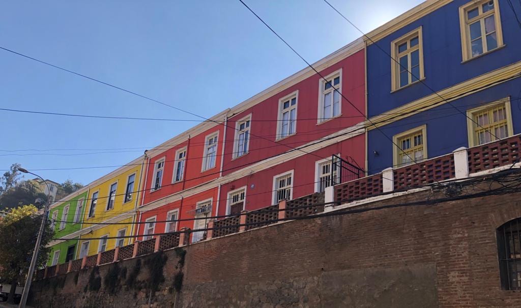 A vibrant building facade presents a colorful display of red, blue, green, and yellow hues. The structure is characterized by large windows framed in white, accented by decorative railings. The lower level features a rustic stone wall, adding texture to the scene.