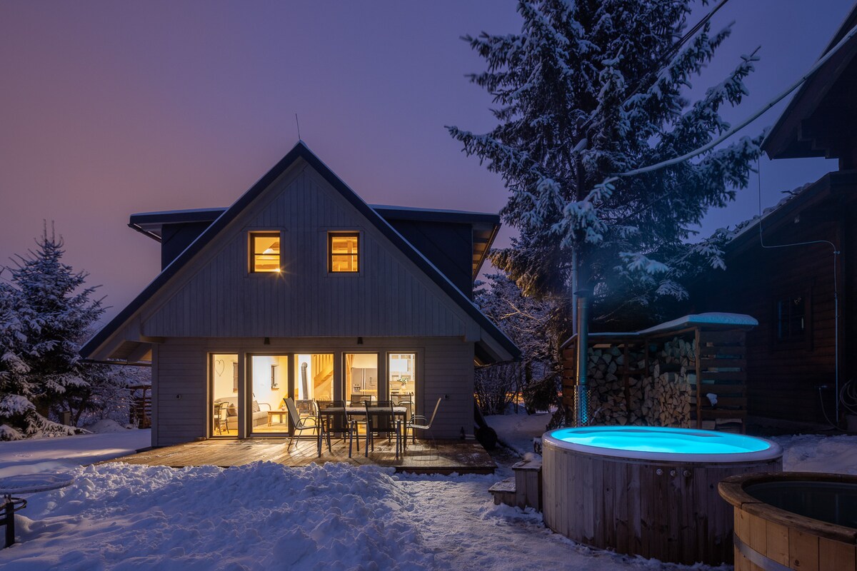 A modern wooden cabin is shown against a twilight sky, with warmly lit windows. An inviting outdoor hot tub sits nearby on a snow-covered deck, surrounded by evergreen trees. The spacious patio features a dining table, enhancing the outdoor experience.