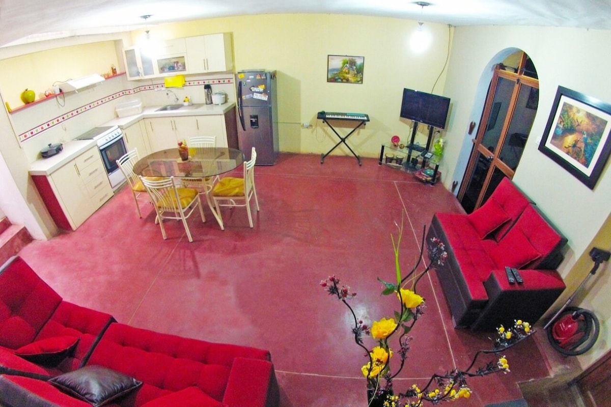 An open-plan kitchen and dining area is featured, equipped with a gas stove, refrigerator, and microwave. A round dining table with white chairs is positioned near the kitchen. Red sofas provide seating in the living area, while a flat-screen TV and keyboard are visible.