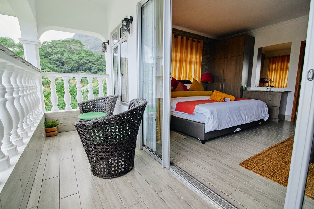 A cozy outdoor seating area is located adjacent to a spacious bedroom. The balcony features two woven chairs and offers views of the surrounding greenery. A brightly colored blanket and pillows accent the bed inside, complemented by warm, flowing curtains that frame the windows.
