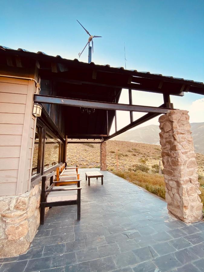An outdoor patio area is featured, showcasing a stone structure with a wooden roof. Two chairs and a small table are positioned on the black stone floor, with an expansive view of the surrounding mountains and wind turbine in the background.