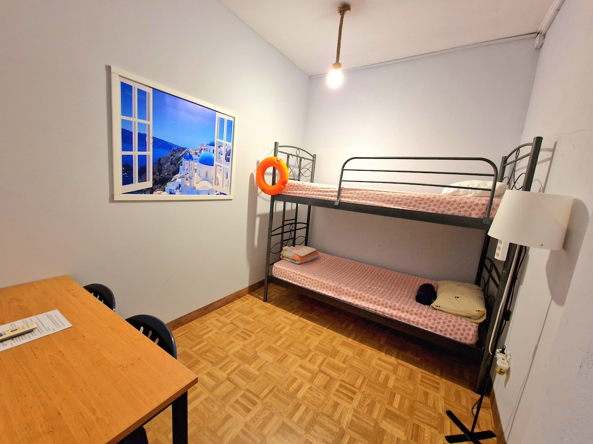 A private room features a metal bunk bed with a pink patterned mattress. A wooden table with black chairs occupies one side, while a light fixture hangs from the ceiling. A framed photograph of a coastal scene decorates the wall, enhancing the simple decor.
