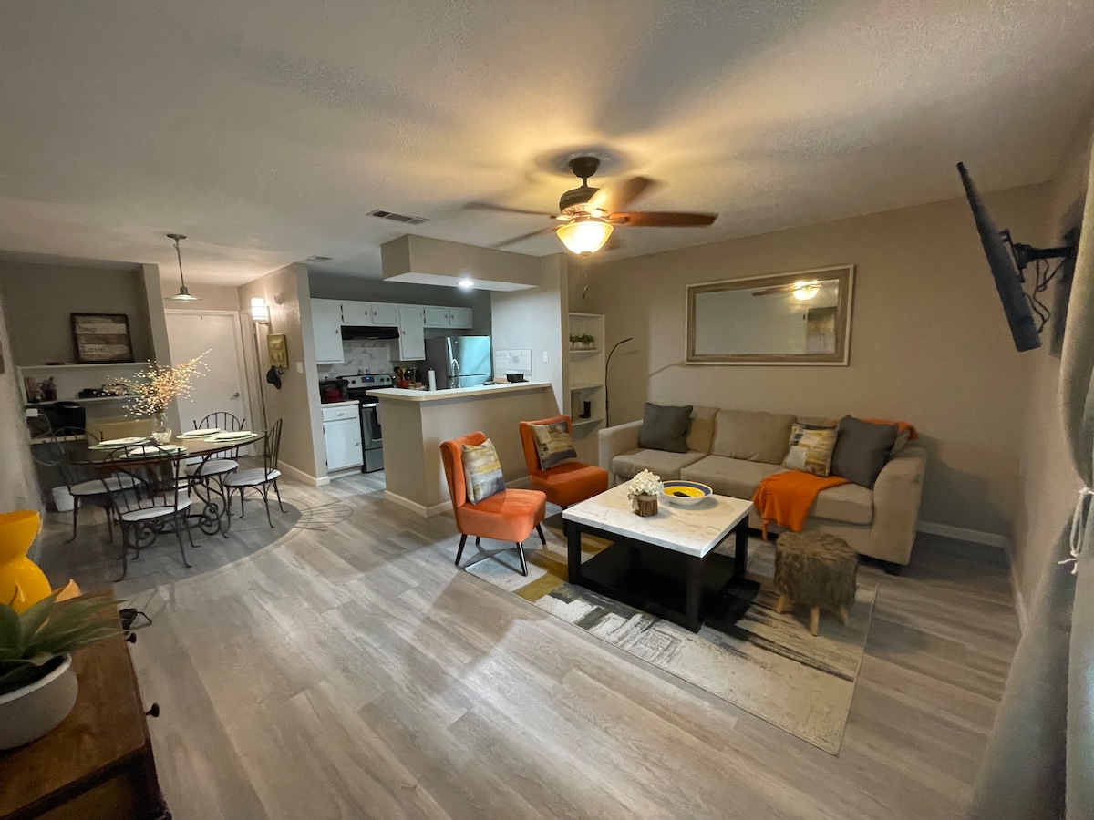 A spacious living area features a comfortable sofa and two vibrant orange chairs, inviting relaxation. A light-toned coffee table sits centrally, with decorative accents. The kitchen area is visible in the background, separated by a counter, while a dining table accommodates four in the adjoining space.