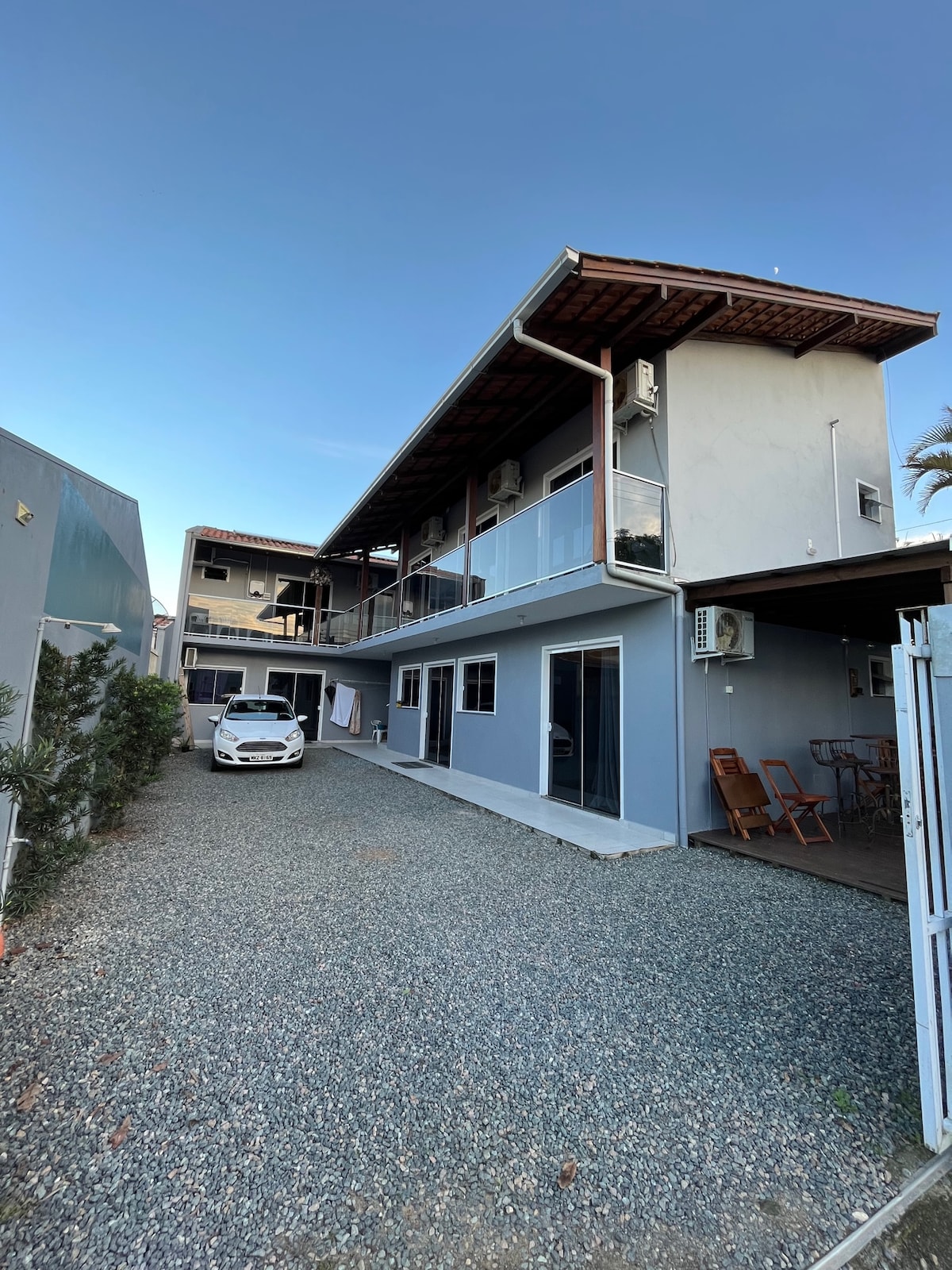 The exterior of a two-story building features a gravel driveway leading to a white car parked near the entrance. Balconies extend from the upper level, while large windows provide ample natural light. The surrounding area includes small shrubs and an outdoor seating arrangement.