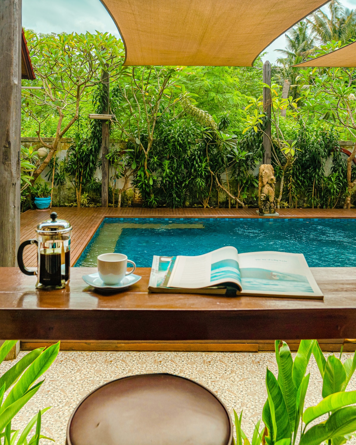 A tranquil corner is showcased, featuring a coffee cup and a French press next to an open magazine. The view extends to a serene pool surrounded by lush greenery and shaded by overhead canopies, creating a peaceful atmosphere.
