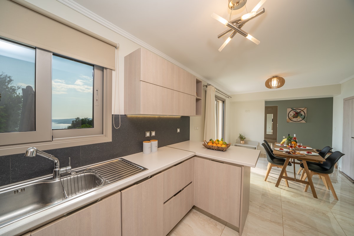 A modern kitchen is presented, featuring light wood cabinetry and a stainless steel sink. Natural light enters through a window, providing a view of the outdoors. Adjacent to the kitchen, a dining area is visible with a table set for meals, surrounded by stylish chairs.