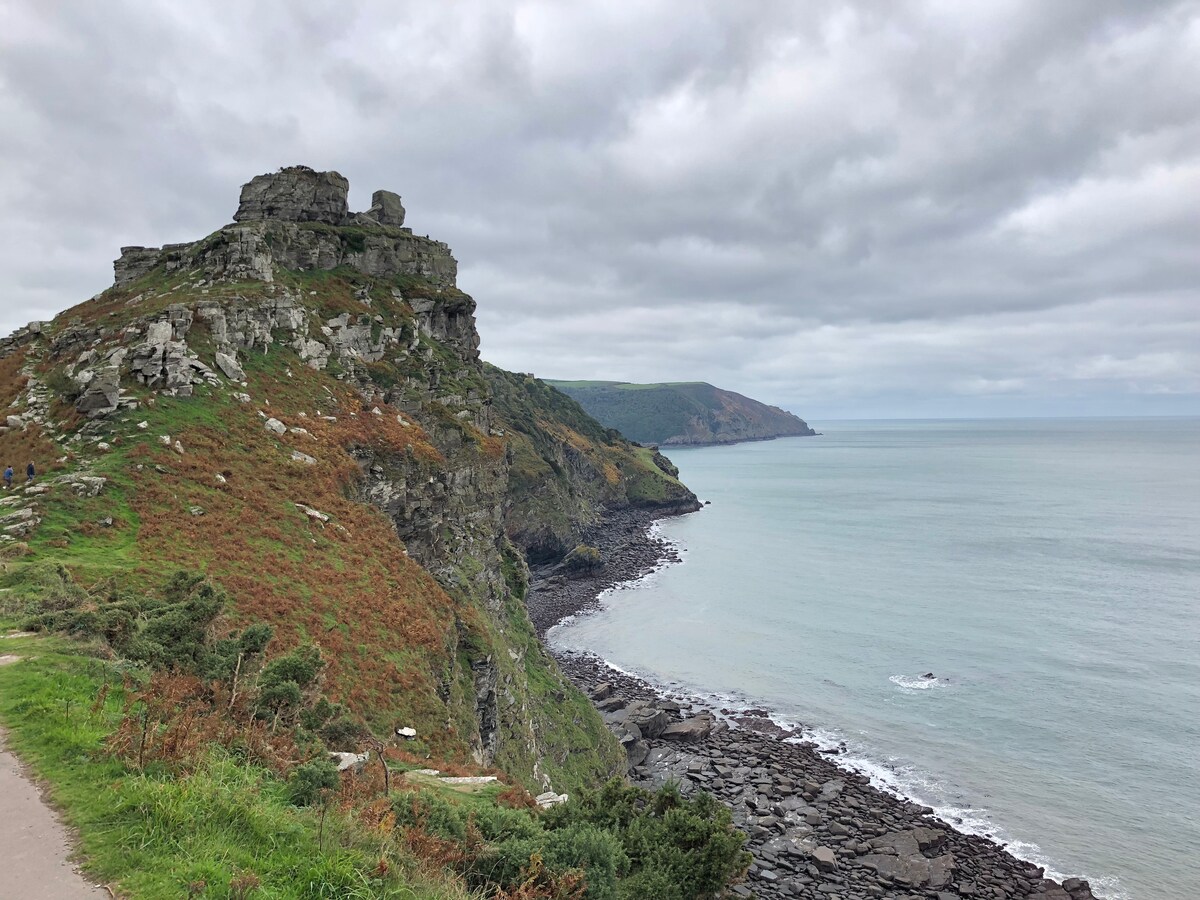 A rugged coastal landscape is captured, featuring a rocky cliff topped with large boulders. The shoreline leads into the calm sea, with scattered rocks visible along the water's edge. Clouds cover the sky, casting soft light over the scene.