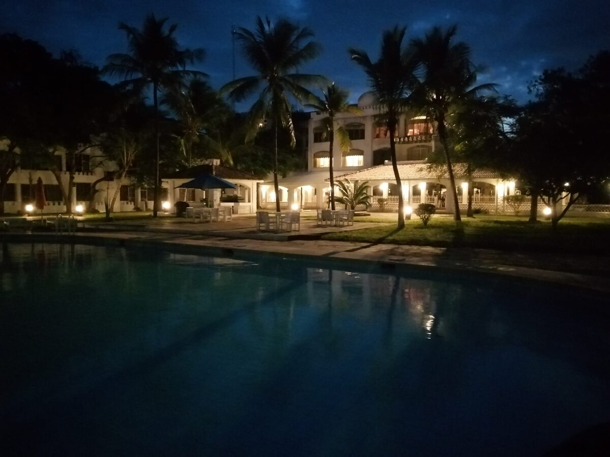 A spacious outdoor area is illuminated at dusk, featuring a large swimming pool surrounded by palm trees. Soft lighting highlights the adjacent buildings, showcasing their architectural details and inviting atmosphere. The serene surroundings contribute to a relaxing ambiance.