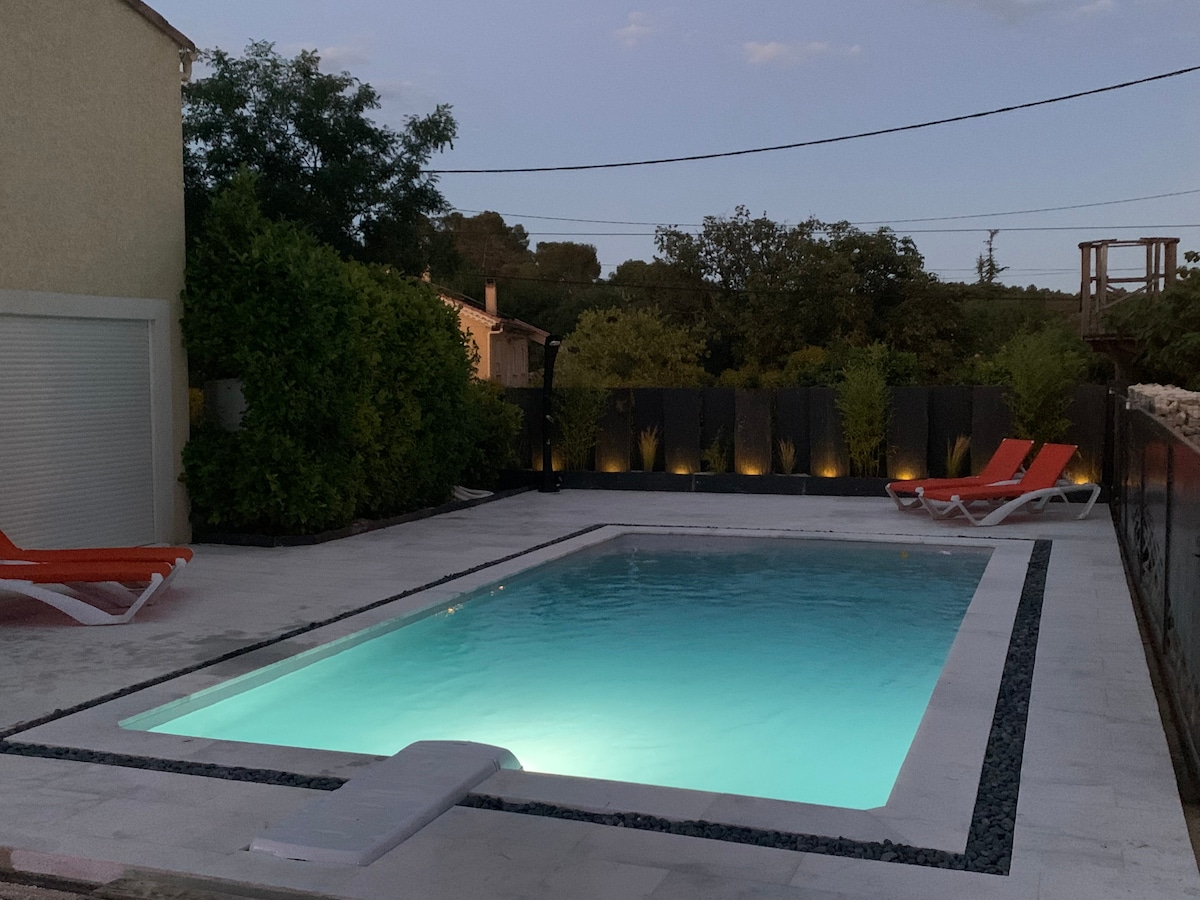 A rectangular swimming pool is set in a spacious outdoor area, illuminated gently by soft lighting along the perimeter. Orange lounge chairs are placed nearby on a clean, tiled surface, surrounded by lush greenery and a tranquil ambiance.