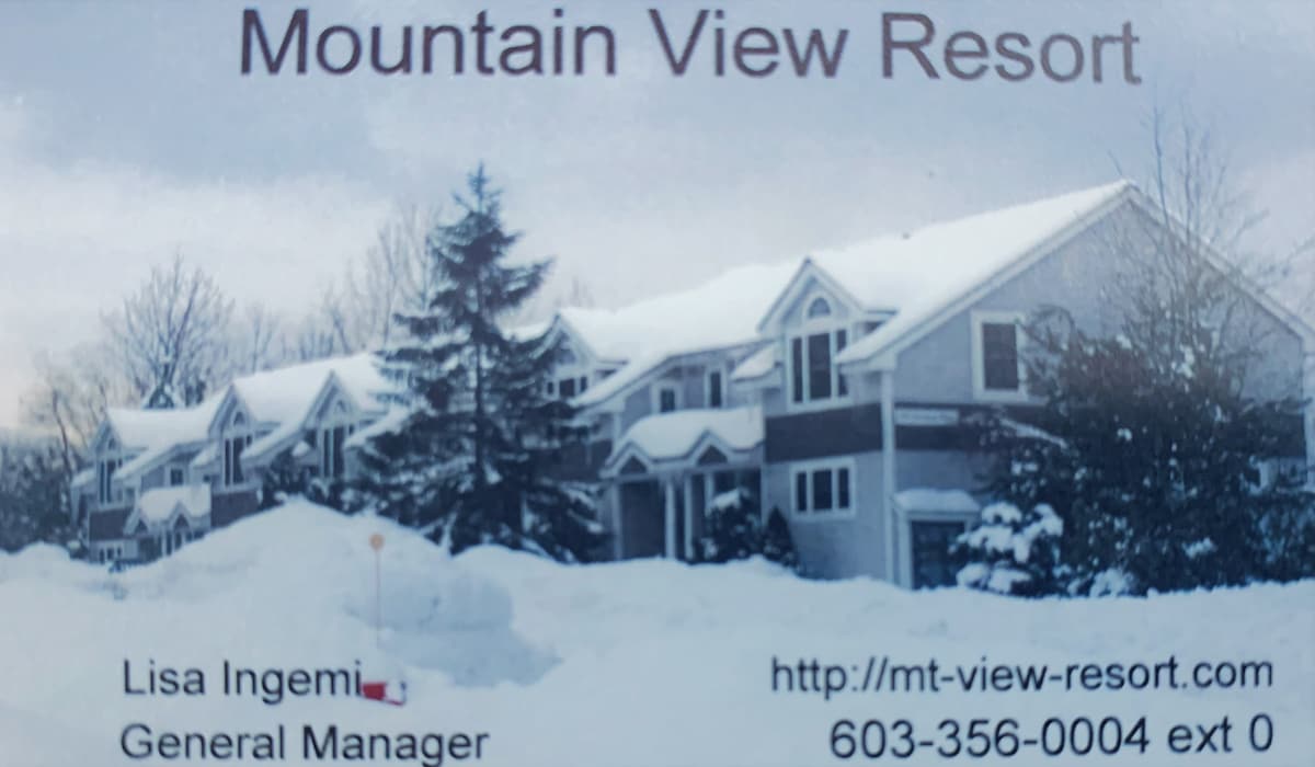 A winter scene showcasing the exterior of the Mountain View Resort, which features multiple two-story buildings with gabled roofs. A thick blanket of snow covers the ground, while evergreen trees frame the resort, creating a serene and snowy landscape.