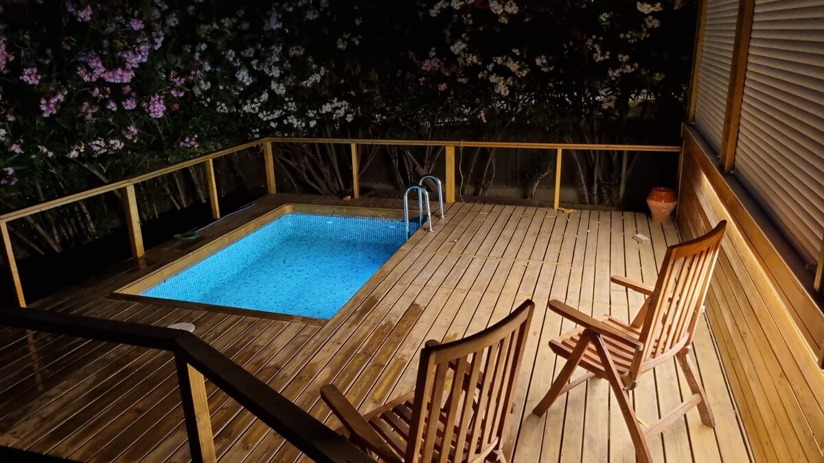 A wooden terrace features a small pool, surrounded by lush greenery. Two wooden chairs are placed nearby, creating a serene atmosphere. Soft lighting highlights the pool's water, adding to the tranquil setting, ideal for relaxation.