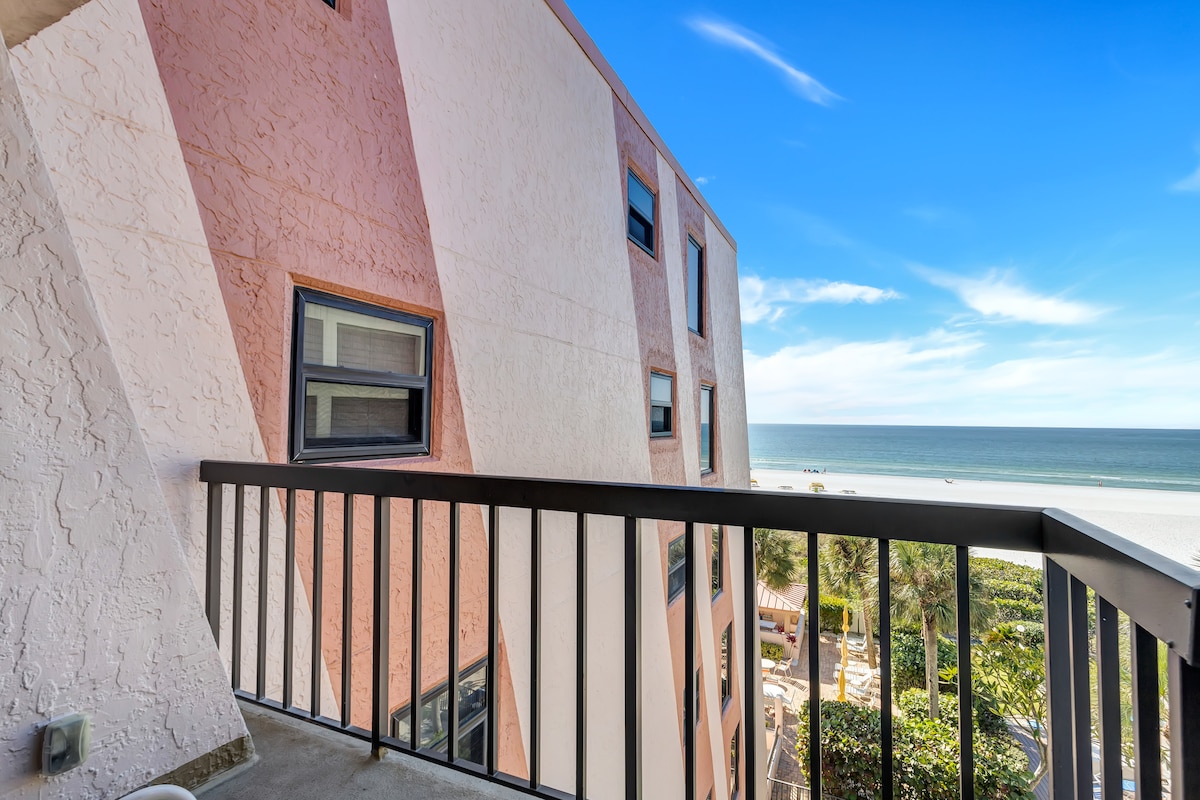 A balcony view showcases the tranquil beach and Gulf waters, framed by the building's pink exterior. The horizon meets the sky, creating an inviting atmosphere for relaxation. Lush greenery is visible below, adding to the serene coastal environment.