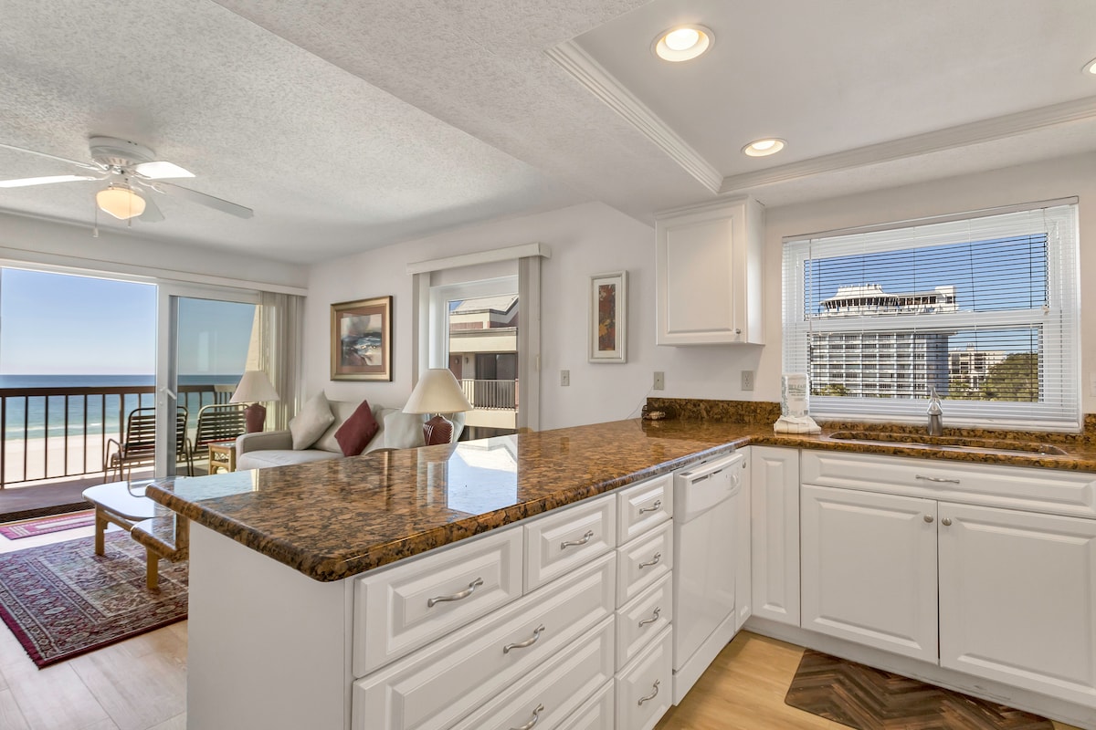 A bright kitchen area features a spacious granite countertop and white cabinetry. Natural light floods the space through a nearby window, showcasing views of the Gulf. The adjacent living area is visible, highlighting a comfortable seating arrangement and access to the balcony.
