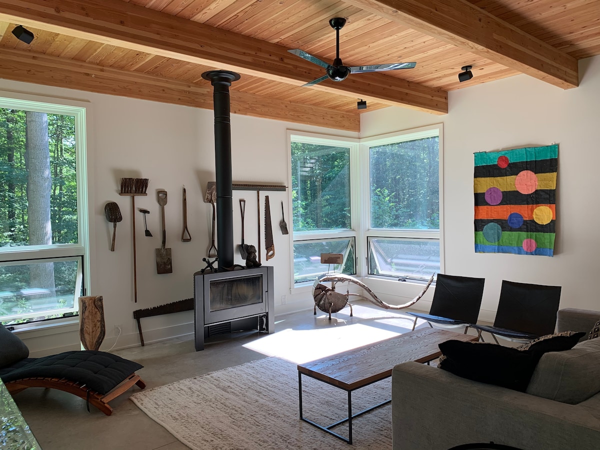 A bright living area features large windows allowing natural light to fill the space. A black wood stove stands beside a minimalist coffee table on a light rug. Cozy seating is provided by a modern couch and two black chairs. A colorful wall hanging adds visual interest.