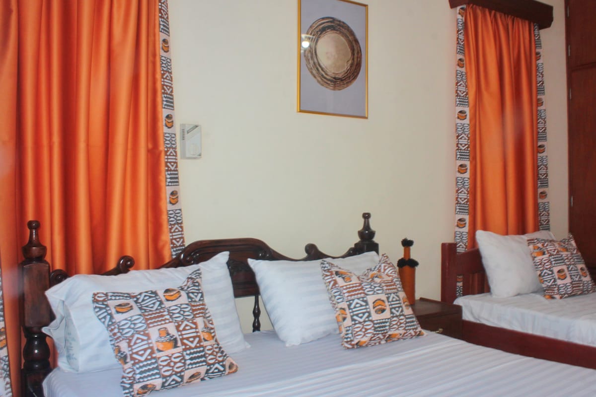 A bright and inviting bedroom features two beds, each adorned with decorative pillows that showcase vibrant patterns. Curtains in warm orange hues frame the windows, complementing the overall color scheme. A piece of wall art hangs above, adding a touch of character to the space.