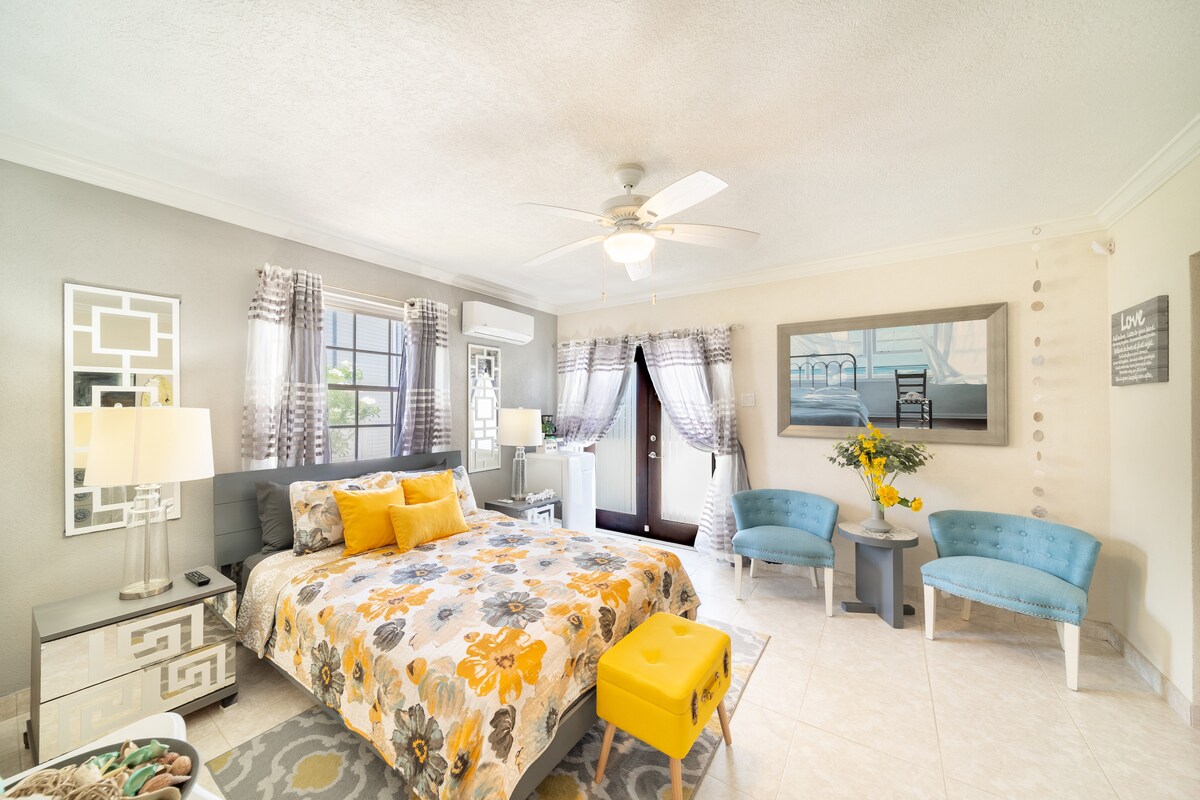 A spacious room features a queen-sized bed covered with a floral comforter, complemented by yellow accent pillows. Two light blue chairs are positioned near large windows adorned with sheer curtains, allowing natural light to fill the space. A decorative floral arrangement adds a touch of color.