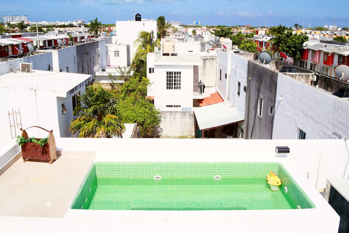 A rooftop terrace features a green-tiled jacuzzi, providing a private space for relaxation. Surrounding views showcase local buildings and rooftops, with a clear sky visible in the background. The area is designed with minimal decoration, emphasizing the pool and scenery.