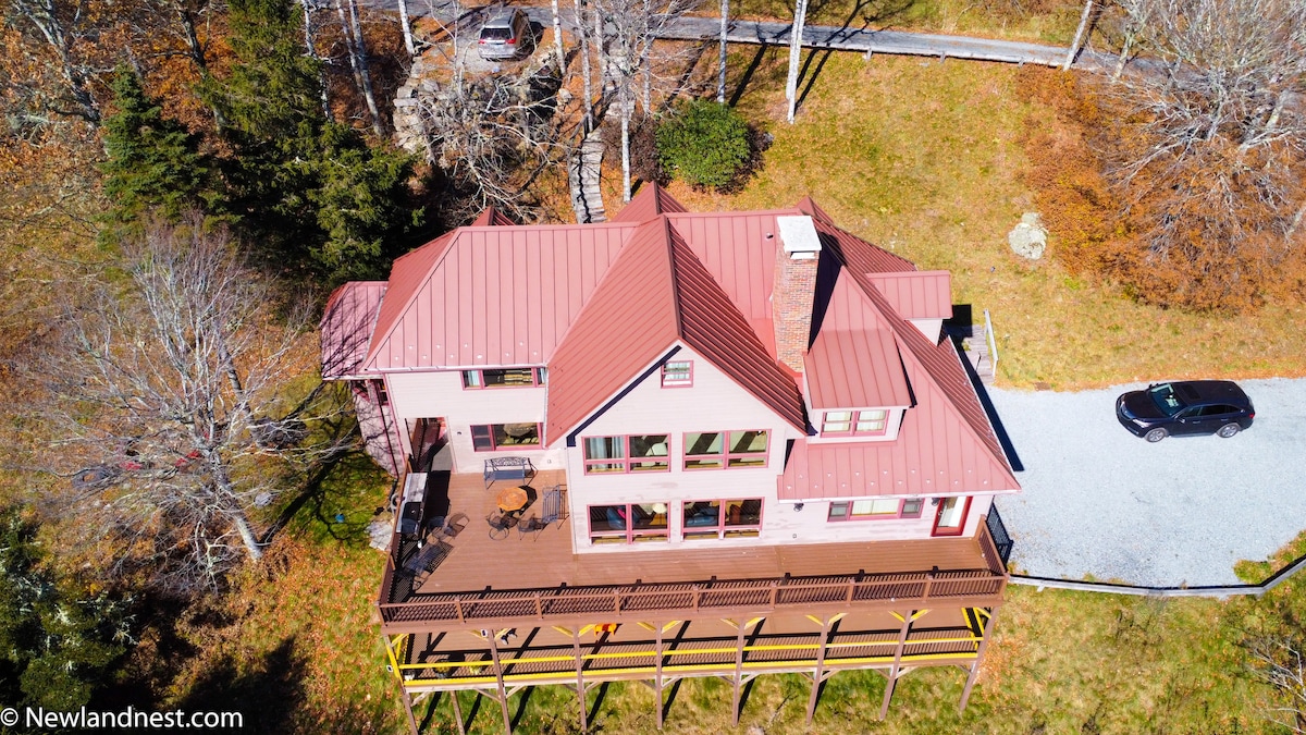 An aerial view of a large, multi-level house set on a grassy landscape. A wooden deck wraps around the structure, and a gravel parking area is visible nearby. Surrounding trees enhance the secluded, tranquil setting.