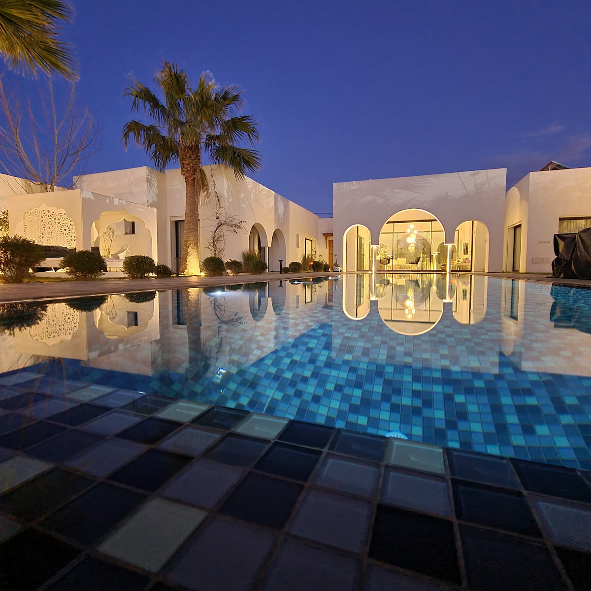 A serene view of the house is reflected in the tranquil pool water at twilight. Palm trees frame the scene, and the structure's elegant archways create a welcoming ambiance. The outdoor space is designed for relaxation, inviting guests to enjoy the calm surroundings.