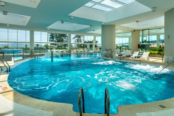 An indoor pool area features a large, circular pool with bubbling water. Large windows provide natural light and views of the surrounding greenery. Several lounge chairs are arranged along the perimeter, creating a spacious and relaxing environment.