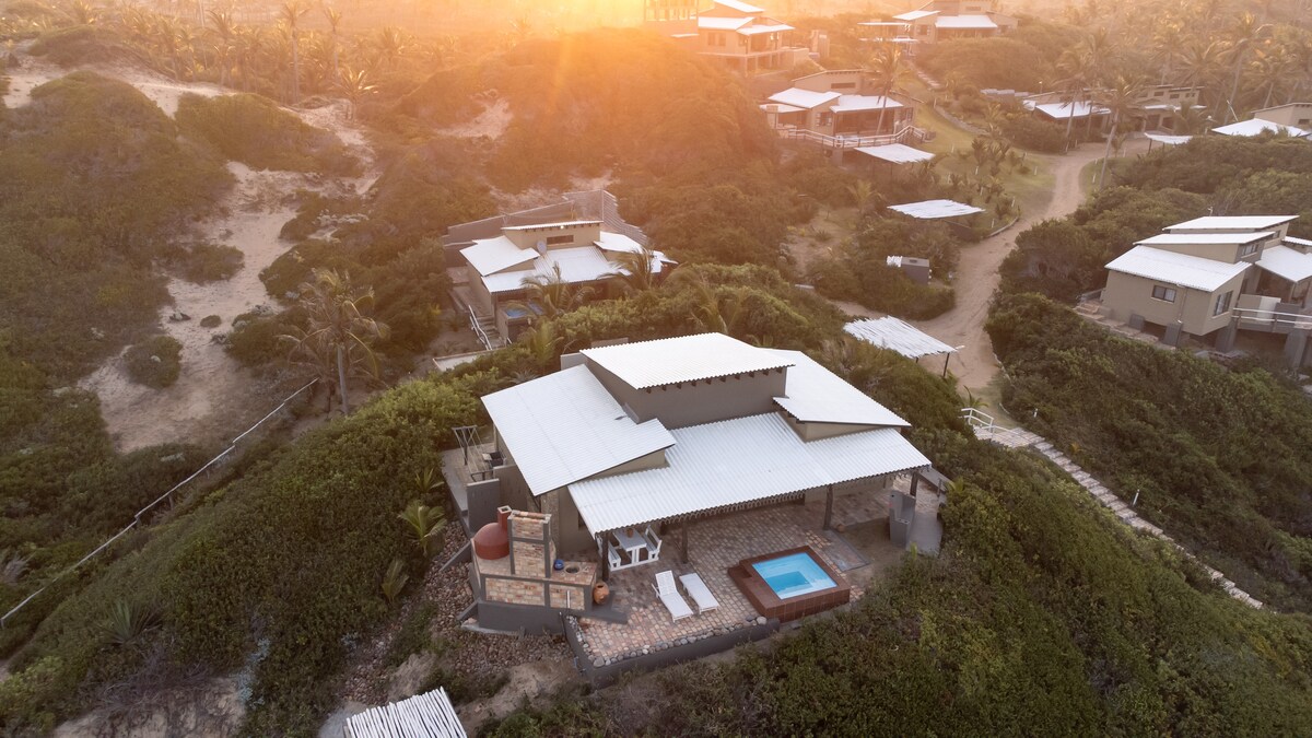 An aerial view captures the lodge nestled among lush greenery, with a private pool and outdoor seating area. The sun sets in the background, casting a warm glow over the villas, which are spaced comfortably apart to ensure privacy.