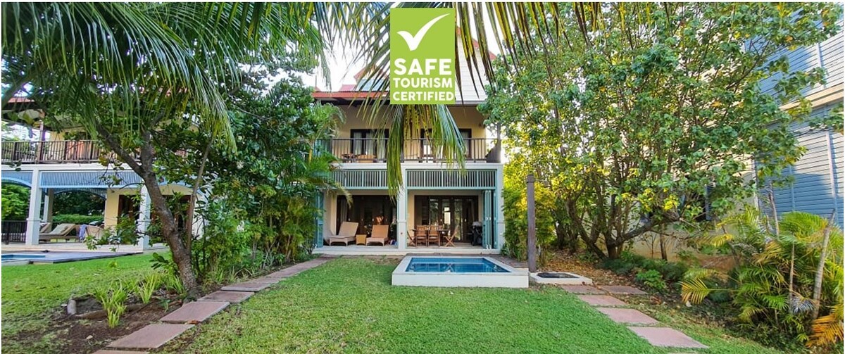 A two-story residence is framed by lush greenery, featuring a covered terrace above an open living space. A small splash pool is located to the side, surrounded by manicured grass and paved pathways, creating an inviting outdoor setting.