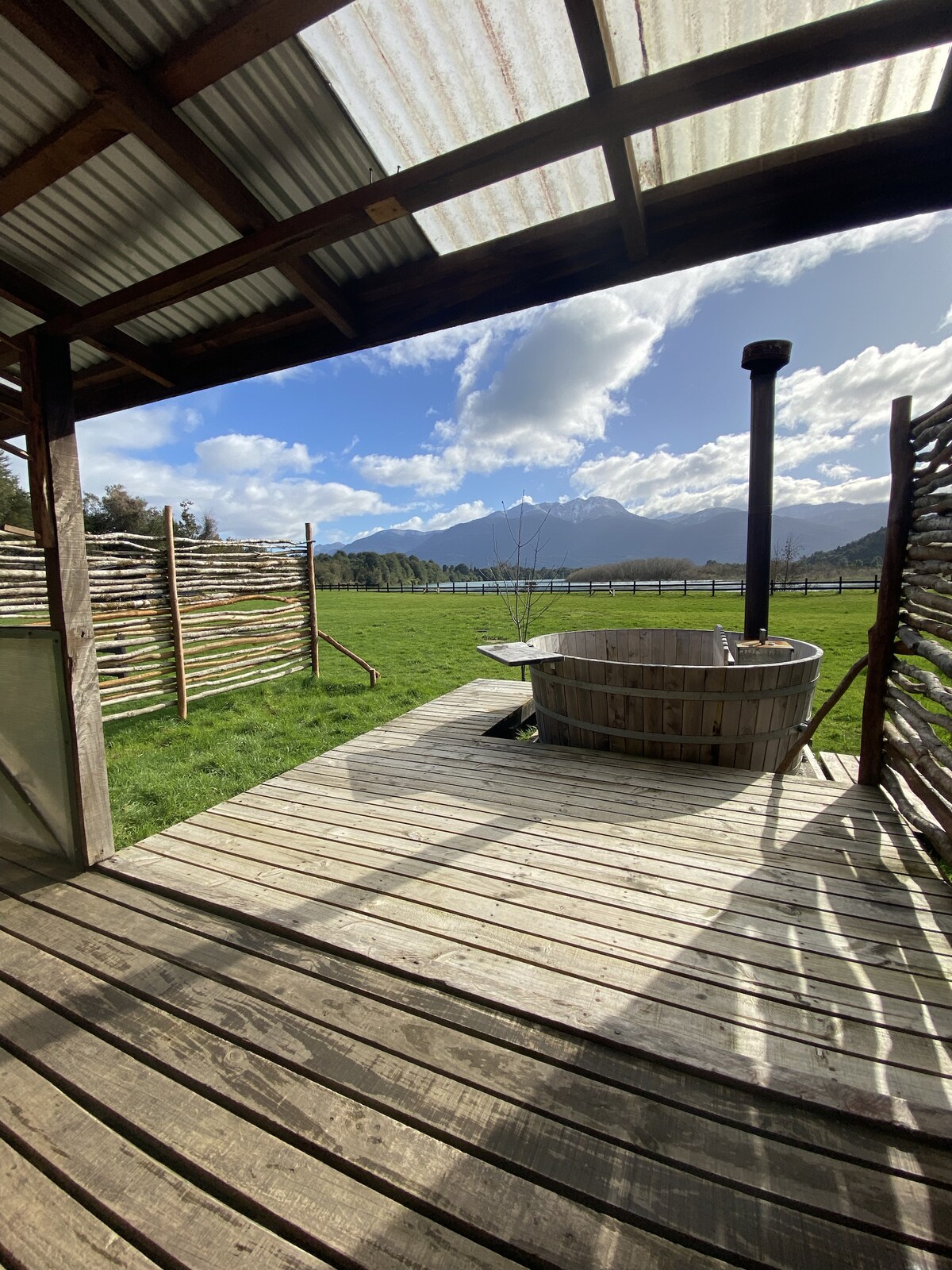 A wooden deck extends from the structure, showcasing a round wooden hot tub surrounded by natural fencing. The expansive green grass and distant mountains can be seen under a partly cloudy sky, with the shimmering river visible in the background.