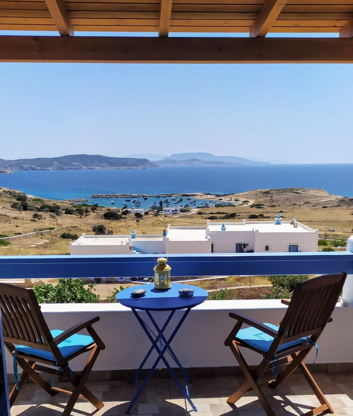 A terrace view showcases a panoramic landscape of the sea and surrounding islands, complemented by traditional white buildings below. Two wooden chairs and a small blue table set the scene for relaxation, inviting moments to enjoy the outdoor atmosphere.