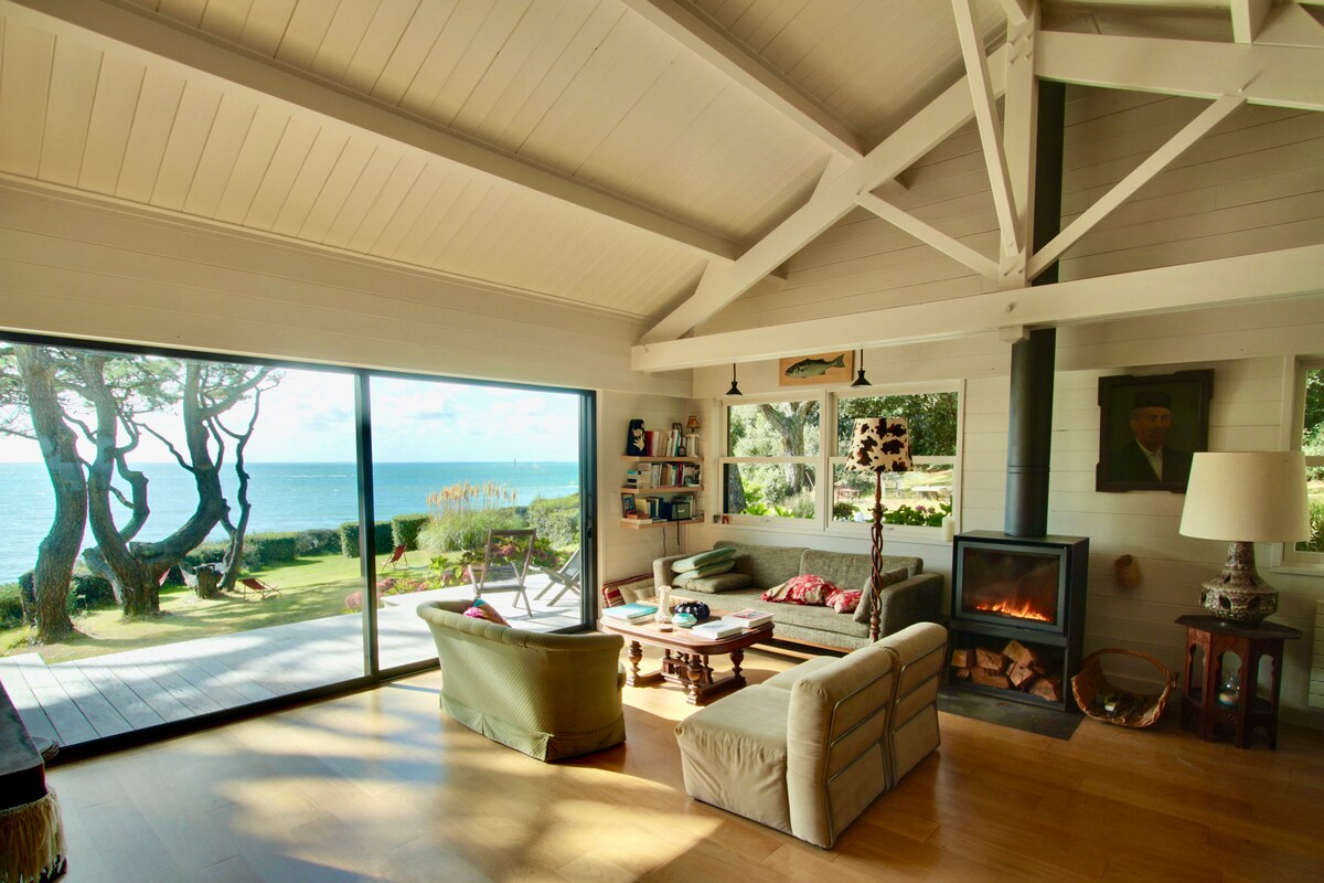 The open living area features large glass doors that offer unobstructed views of the ocean and garden. A cozy seating arrangement is framed by a high ceiling supported by exposed beams. A fireplace adds warmth to the space, enhancing the serene coastal atmosphere.