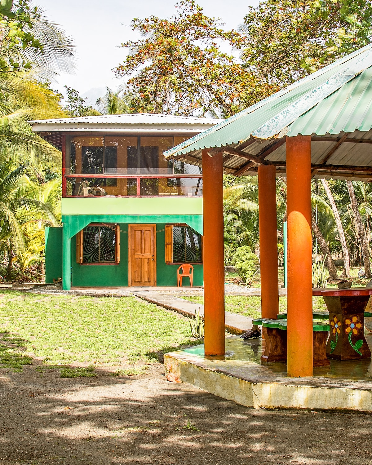 A vibrant two-story cottage stands surrounded by lush greenery and palm trees. The exterior features a green facade with warm wooden accents and large windows. An open-air gazebo with colorful pillars and a rustic table adds a charming outdoor space, inviting relaxation amidst nature.