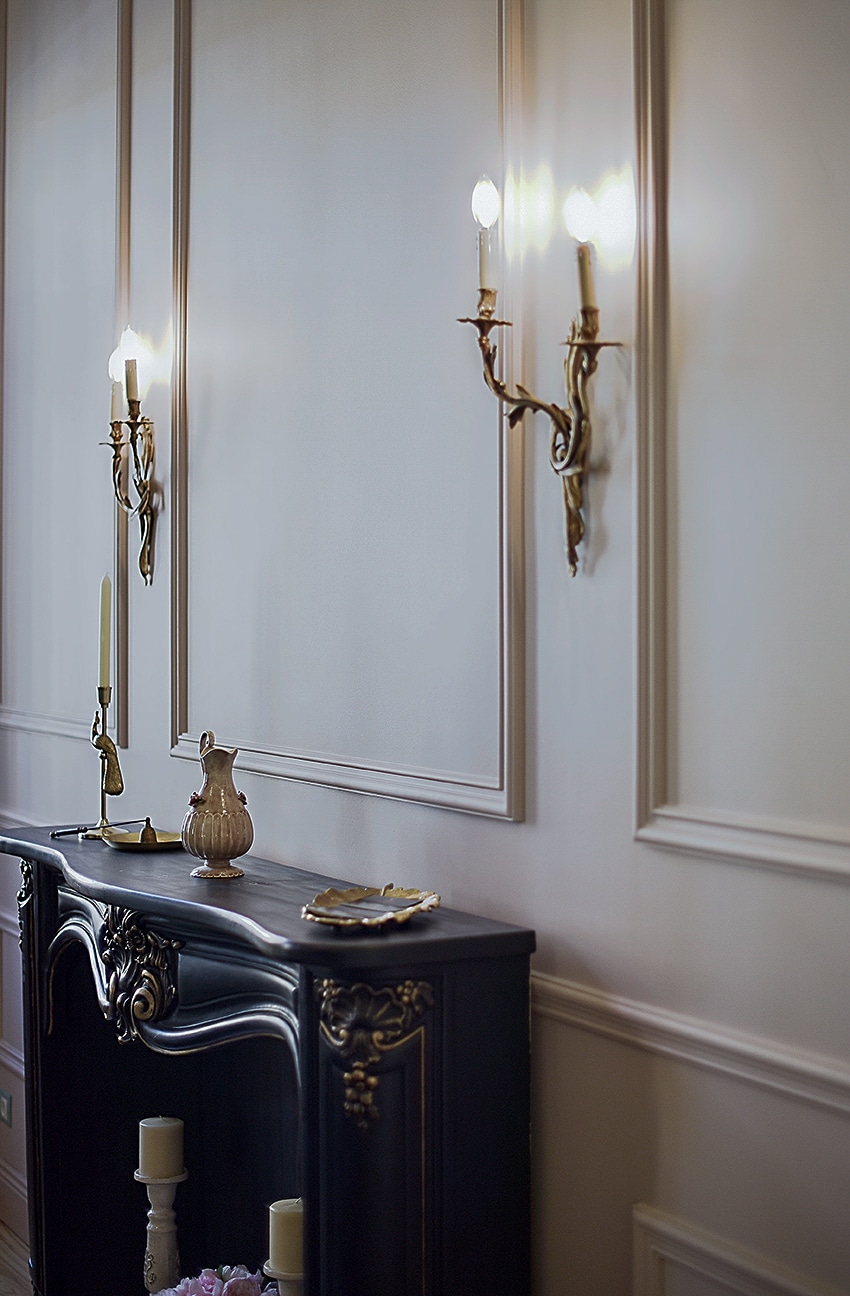 An elegant wall features two antique-style wall sconces, casting a warm glow. A dark wooden console table rests beneath, adorned with decorative items and candles. The walls display intricate moldings, enhancing the refined ambiance of the space.