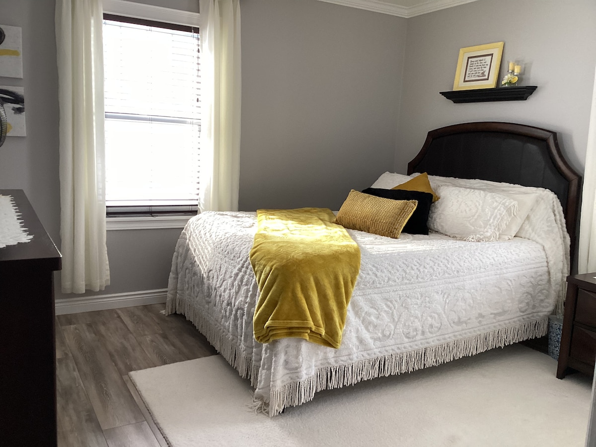 A private bedroom is presented with light grey walls and a large window dressed in flowing light curtains. A spacious bed is adorned with a white patterned coverlet and a bright yellow throw blanket. A dresser and artwork add to the welcoming decor.
