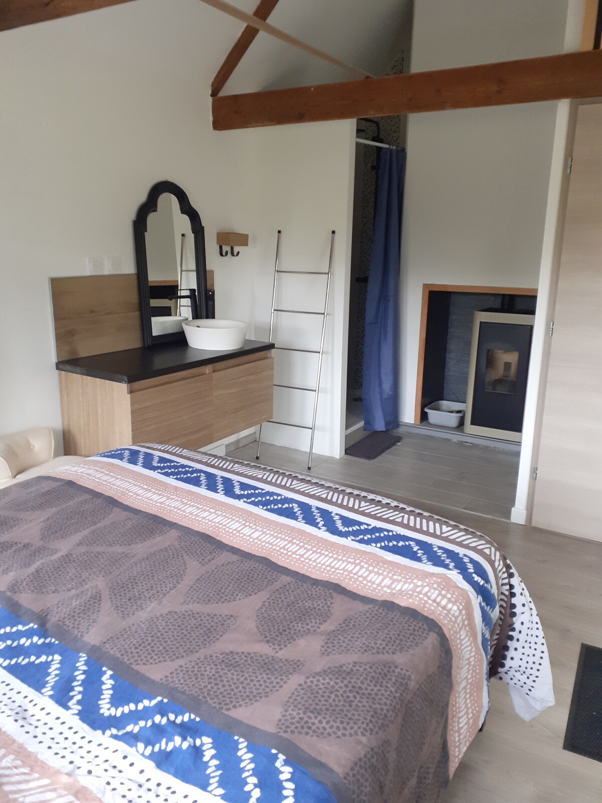 A cozy room features a spacious bed with a patterned blanket. A wooden dresser with a mirror is positioned nearby, alongside a ladder shelf. A shower area is enclosed by a curtain, and a small fireplace is visible in the corner, adding warmth to the space.