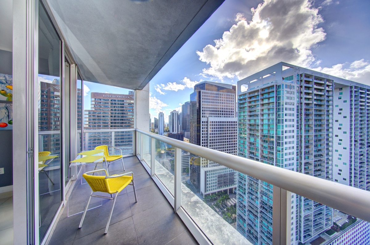 A spacious balcony features two yellow chairs positioned against a glass railing, offering clear views of the city skyline. The scene captures a mix of modern buildings and a bright sky with scattered clouds, creating a vibrant urban atmosphere.