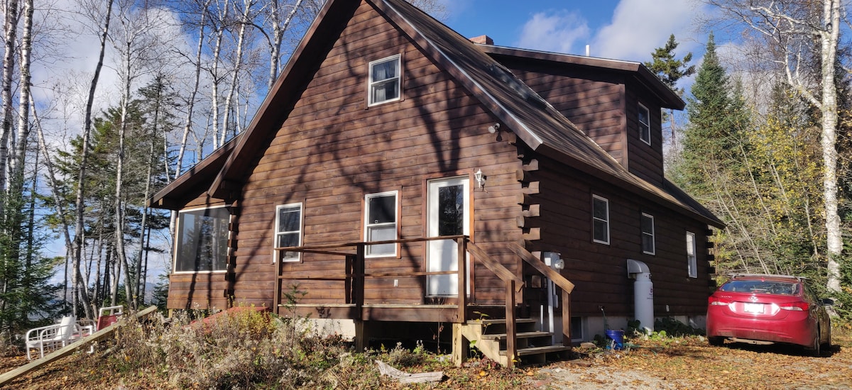 The log house exterior features a rich brown wood façade, adapted to its natural surroundings. A spacious deck with steps provides access to the entrance. Trees surround the property, adding privacy, while a vehicle is parked nearby on the gravel driveway.
