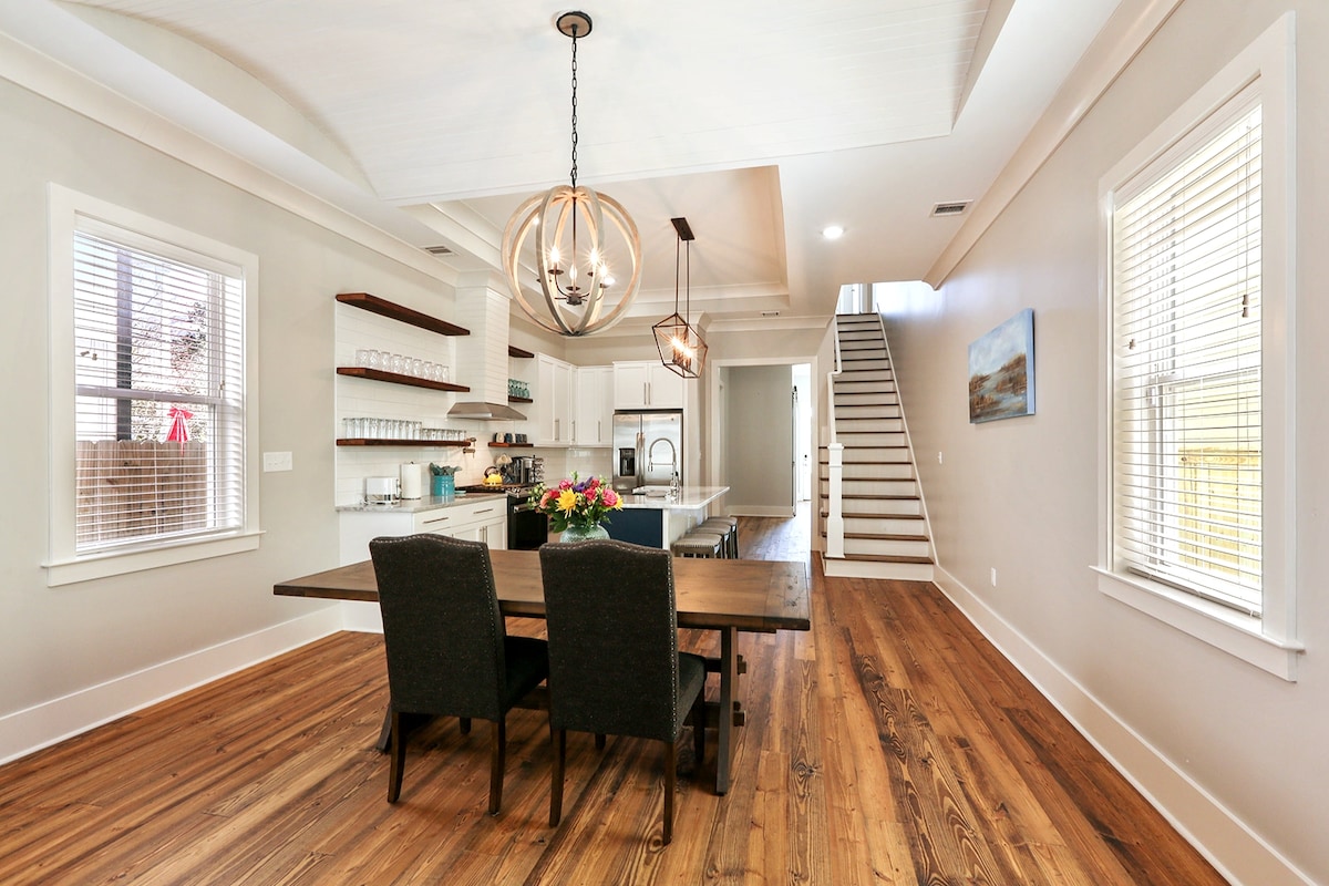 An open dining area is featured with a wooden table, accompanied by two upholstered chairs. Natural light enters through multiple windows, illuminating the space. In the background, a modern kitchen is visible, along with a staircase leading to the upper level.
