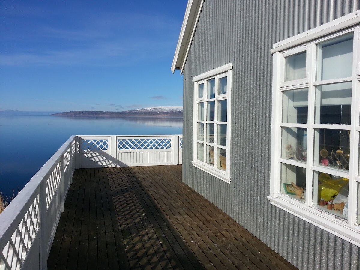 The exterior of a gray, corrugated house is viewed from an expansive wooden terrace. Large windows allow for scenic views of a calm body of water, while reflections of the surrounding landscape can be seen. The terrace railing features a decorative white lattice design.