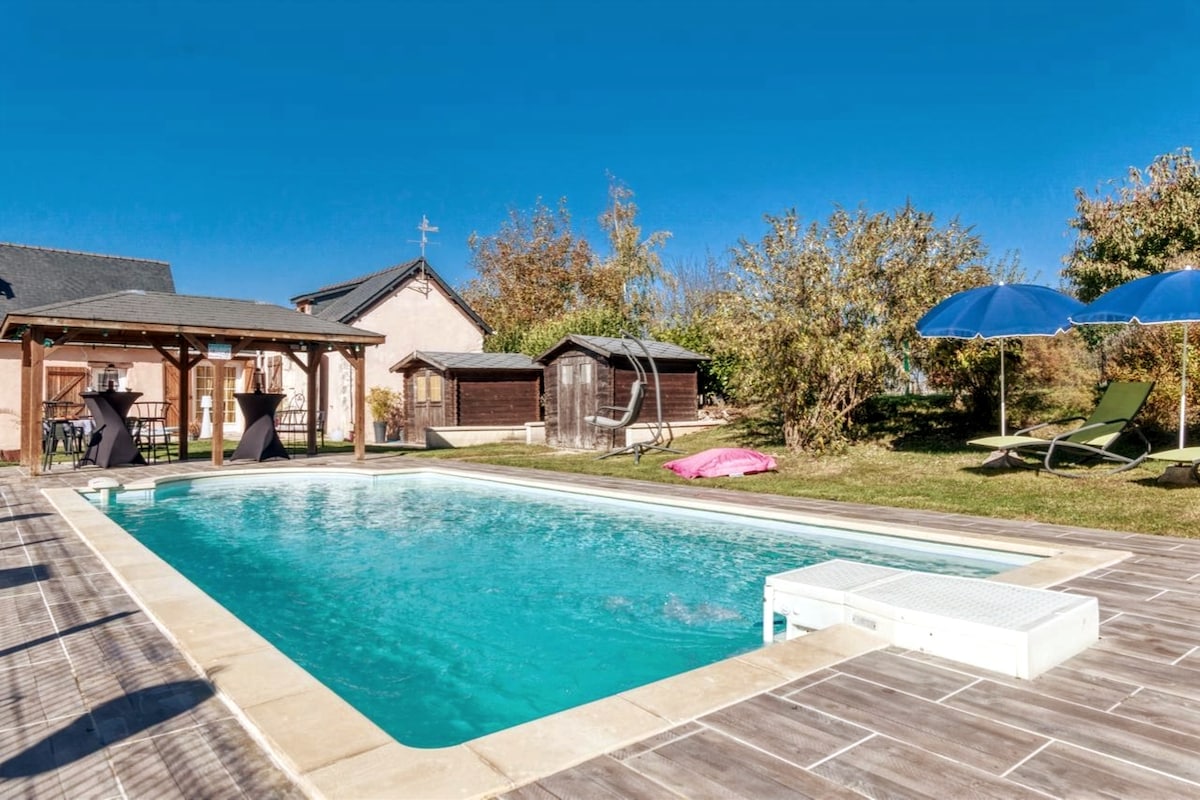 A private swimming pool is surrounded by a sunbathing area featuring umbrellas and lounge chairs. Nearby, several sheds are visible, along with a grassy area dotted with trees, offering a peaceful outdoor setting.