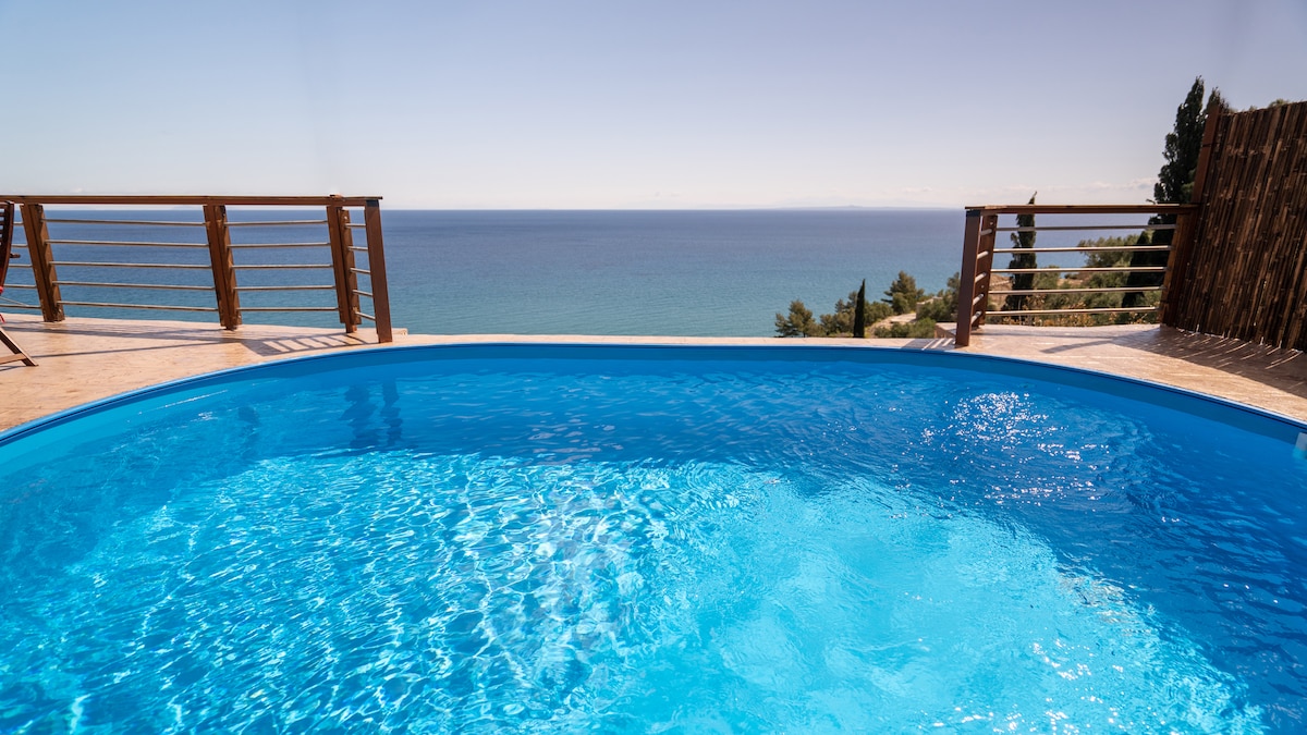 A circular pool features clear, shimmering water, set against a coastal backdrop. Wooden decking surrounds the pool, offering unobstructed views of the sea and a bright blue sky. The serene atmosphere is complemented by the lush greenery in the distance.