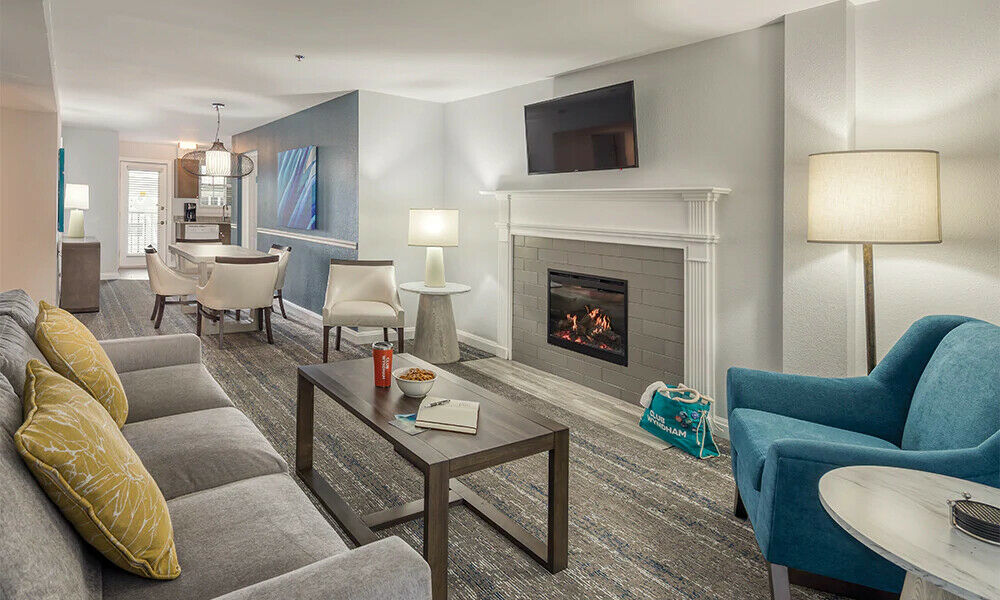 A well-appointed living area includes a comfortable gray sofa with yellow accent pillows, a modern coffee table, and two stylish chairs. A flat-screen TV hangs above a cozy fireplace. Soft lighting is provided by table lamps, creating a warm ambiance.