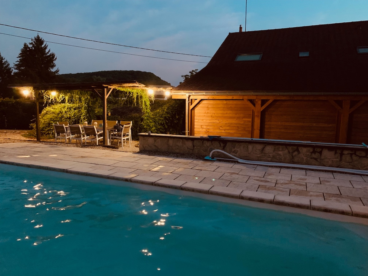 A private pool is set along a paved area, illuminated softly by overhead lights. A shaded dining space with multiple chairs is situated nearby, surrounded by tall greenery. The house features a rustic wooden structure, contributing to the welcoming ambiance of the outdoor setting.