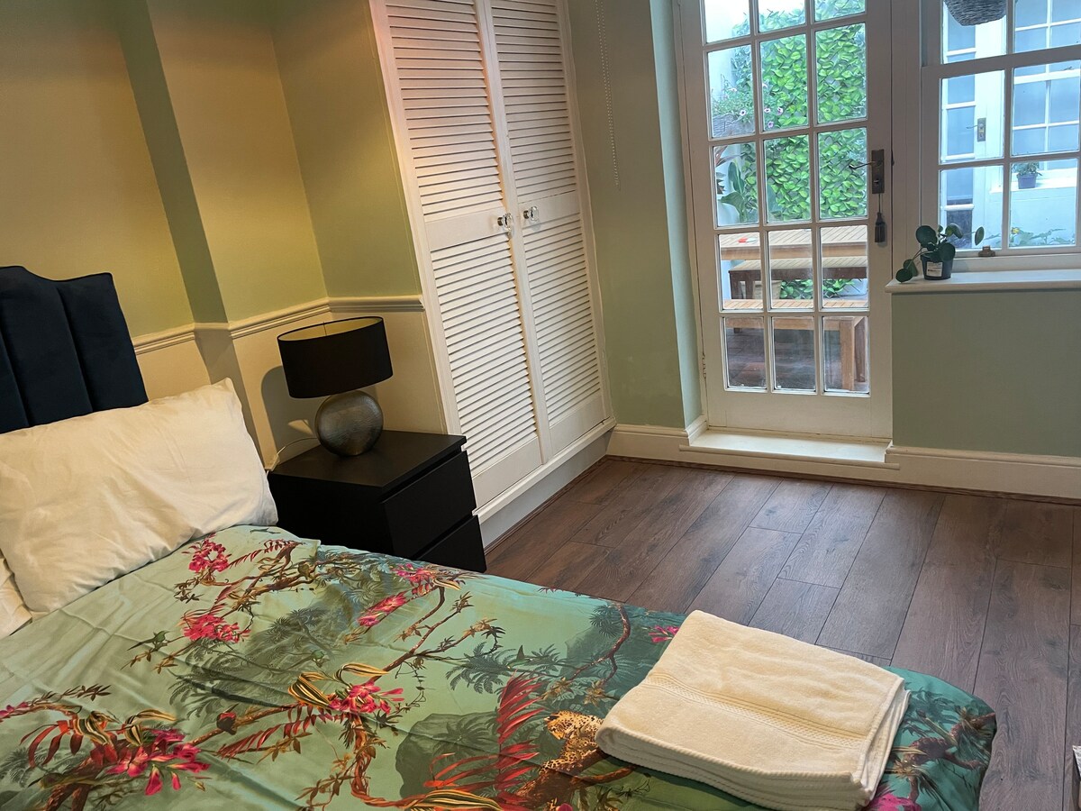 A well-appointed bedroom features a bed with floral bedding and a neatly folded towel. A bedside table with a lamp is positioned next to the bed. Natural light enters through a window, revealing a view of greenery and an outdoor seating area.