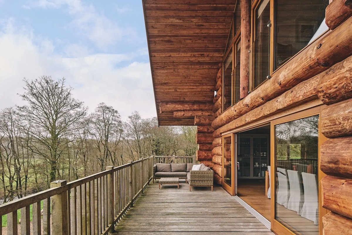 The spacious outdoor deck is framed by rustic log walls, featuring comfortable seating arrangements. Large glass doors invite natural light into the interior, while a scenic view of surrounding trees enhances the peaceful outdoor environment.