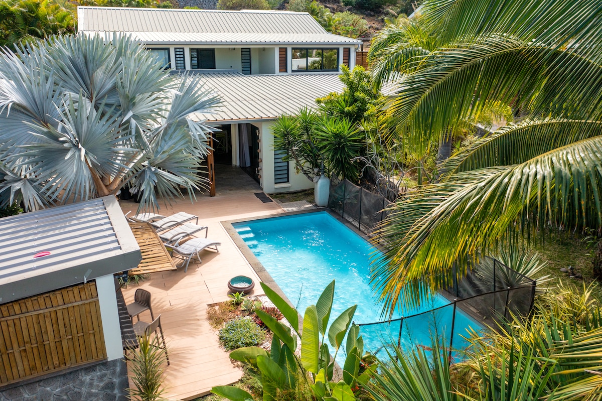 A spacious outdoor area features a private pool surrounded by lush greenery and tropical plants. Sun loungers are positioned near the pool, while a covered space with a wooden structure is visible to the side. The villa's exterior showcases a welcoming entrance and large windows.