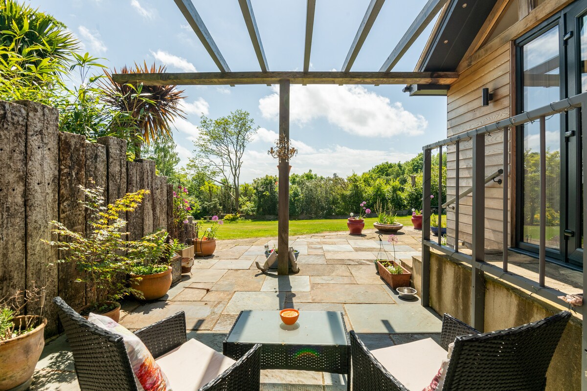 A private patio area is presented with comfortable seating arranged around a small table. Potted plants are placed throughout the space, and a full glass entrance leads to a lush garden area, offering a serene outdoor retreat bathed in natural light.