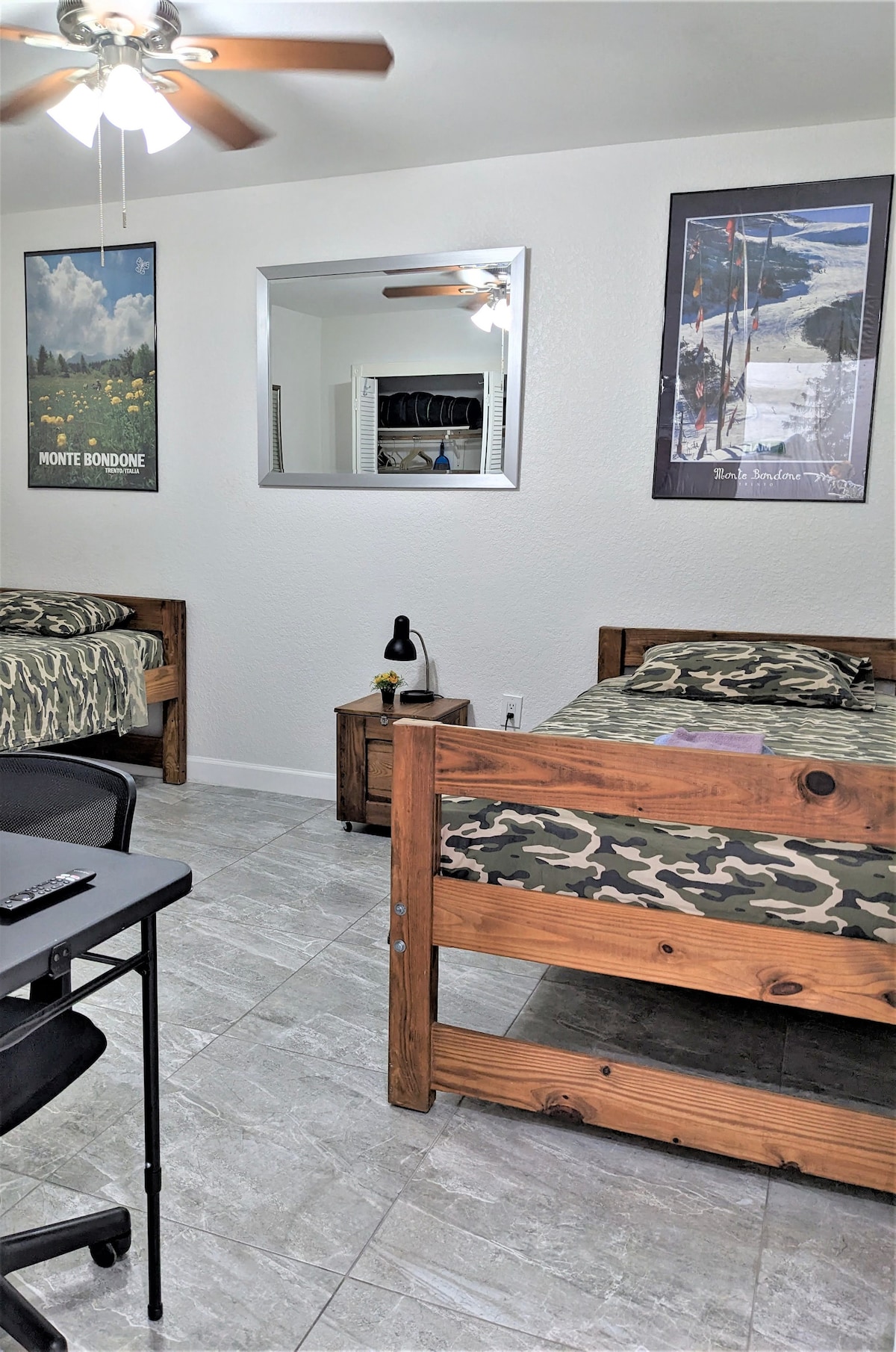 The room features two twin beds with wooden frames, each adorned with camouflage-patterned bedding. A small nightstand is located between the beds, accompanied by a lamp. A large mirror reflects the room, while artwork adorns the walls, contributing to a functional and inviting space.
